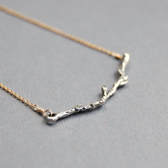 A lightly oxidized sterling silver cast curved twig charm is wire wrapped to the center of a delicate 14k gold filled chain on a gray background.
