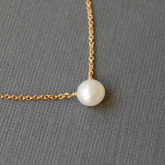 Closeup of a white pearl on a grey background. The pearl is drilled off-center, so it hangs more like a pendant. The pearl is on a delicate 14k gold filled chain.