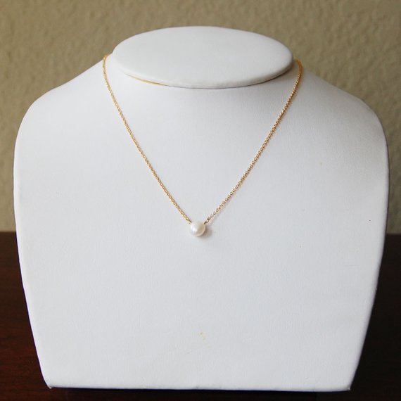 The white pearl solitaire necklace on a delicate gold chain is displayed on a white necklace bust. The pearl is centered at the front of the chain.