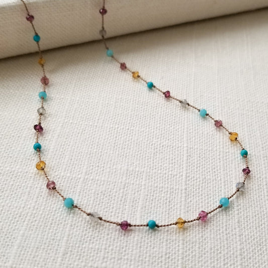 Necklace featuring citrine, labradorite, amazonite, turquoise, pink tourmaline, and garnets evenly spaced, knotted on beige silk cord, is displayed on a white fabric background.