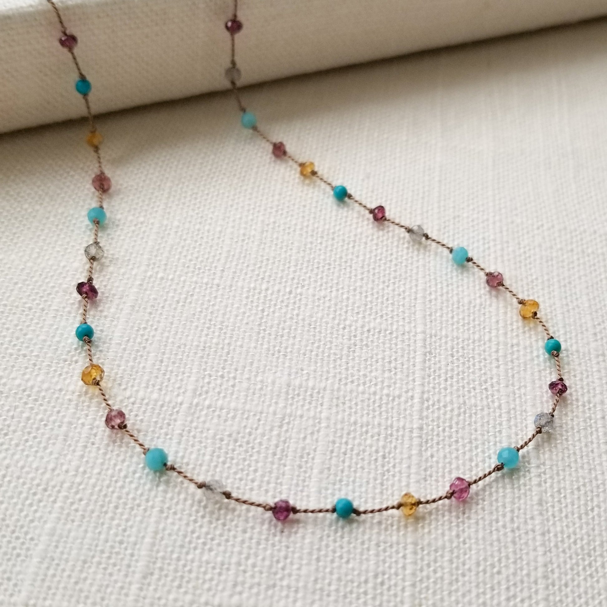 Necklace featuring citrine, labradorite, amazonite, turquoise, pink tourmaline, and garnets evenly spaced, knotted on beige silk cord, is displayed on a white fabric background.