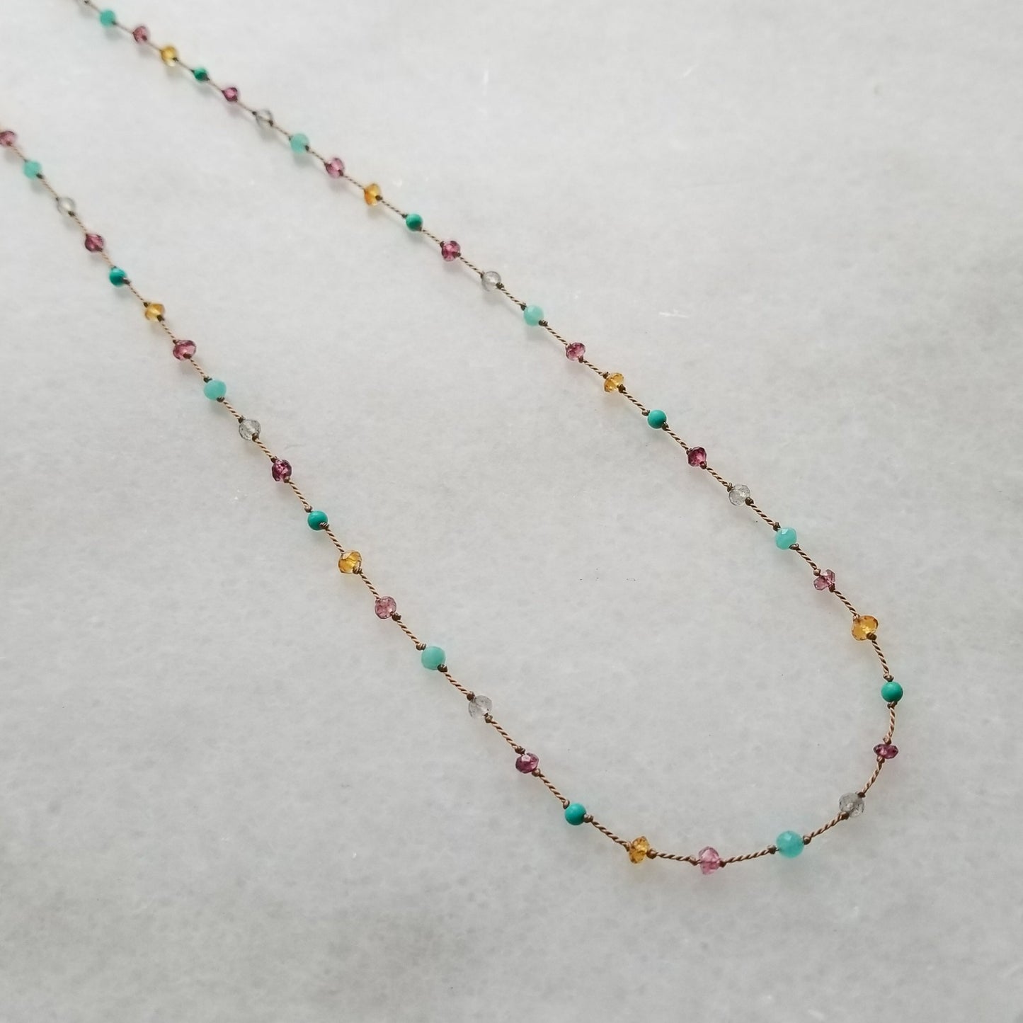 Necklace featuring citrine, labradorite, amazonite, turquoise, pink tourmaline, and garnets evenly spaced, knotted on beige silk cord, is displayed on a white marble background.