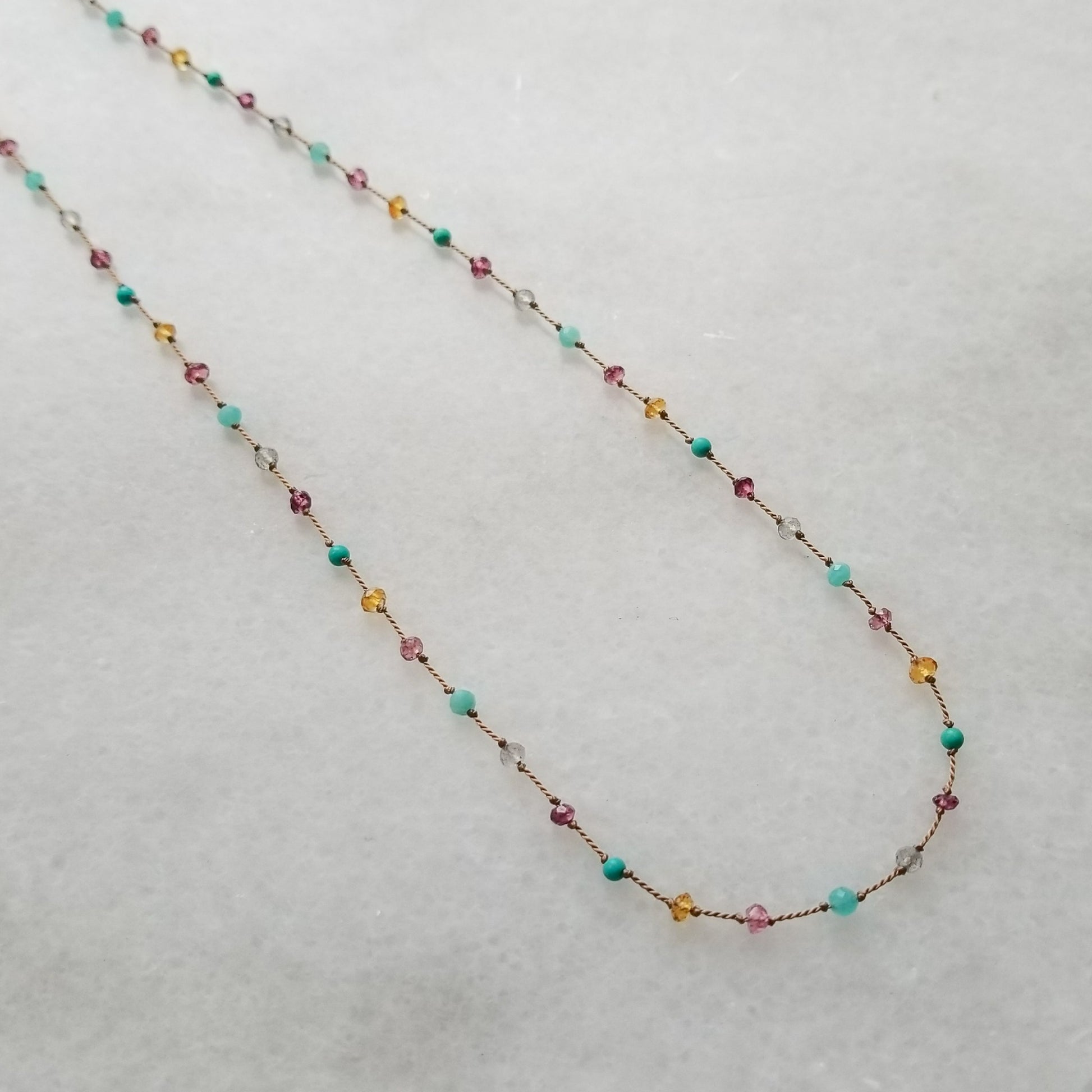 Necklace featuring citrine, labradorite, amazonite, turquoise, pink tourmaline, and garnets evenly spaced, knotted on beige silk cord, is displayed on a white marble background.