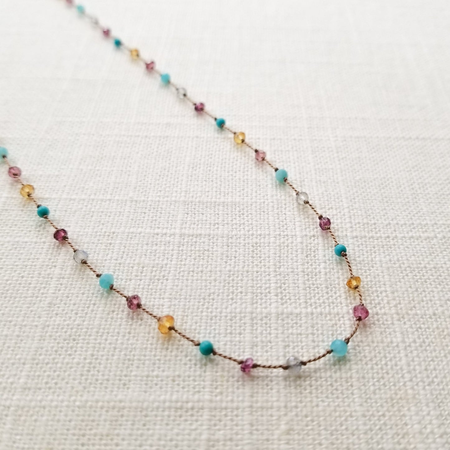 Necklace featuring citrine, labradorite, amazonite, turquoise, pink tourmaline, and garnets evenly spaced, knotted on beige silk cord, is displayed on a white fabric background.